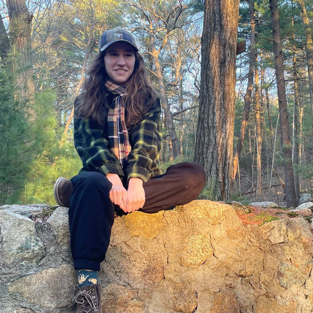 Photo of K.R.Rose sitting on a rock wall in the middle of the woods. She is a young, white person with long, brown hair down to her shoulders. She is wearing a green flannel and black pants. 